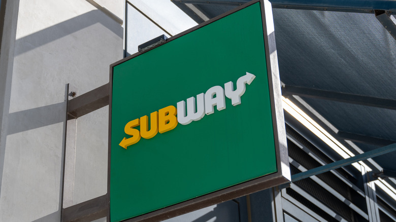Subway restaurant hanging sign with logo on green background attached to a building on a sunny day