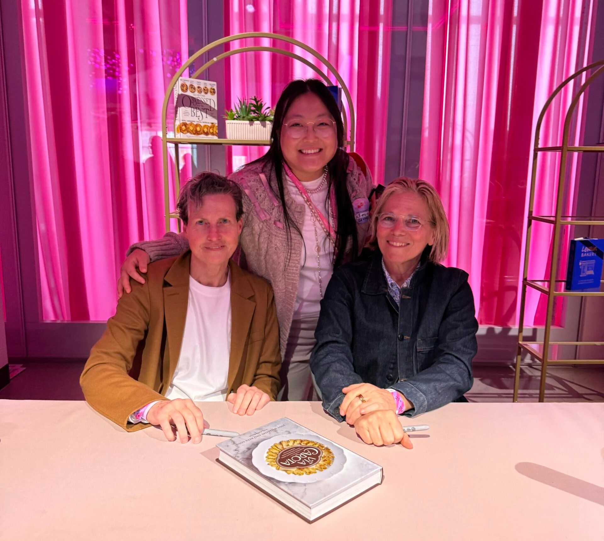 Kaitlin Leung at a book signing with Via Carota chefs Rita Sodi and Jody Williams at a pink table in front of a hot pink wall.