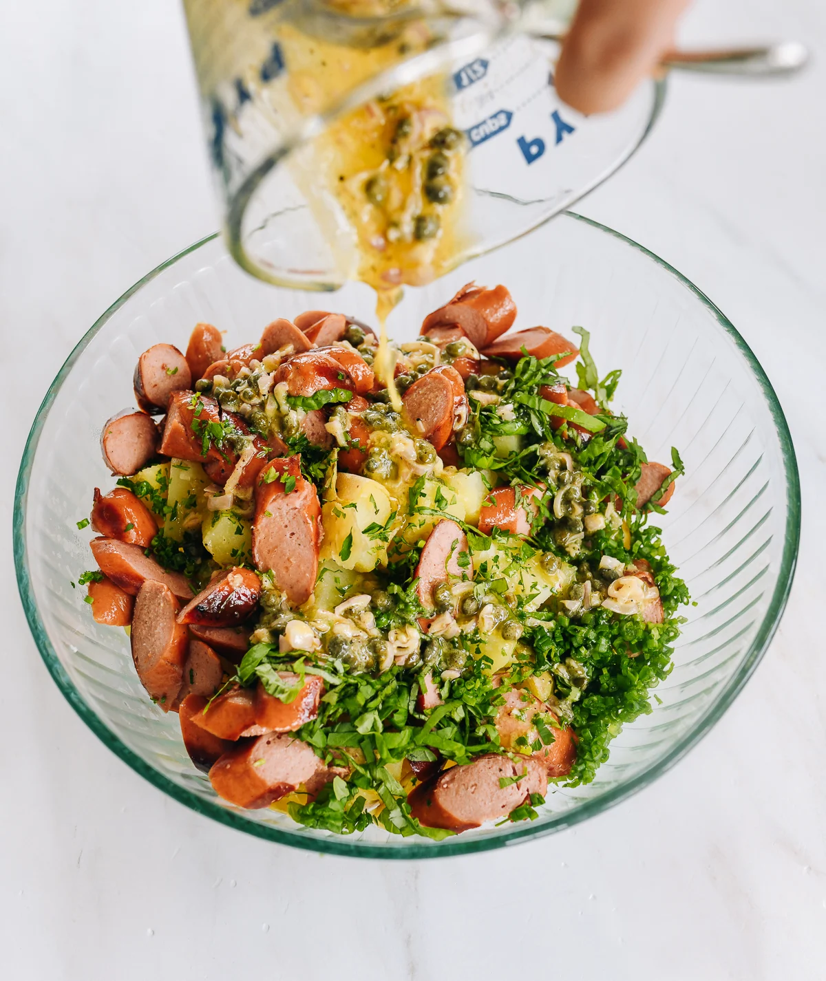 pouring dressing over hot dogs, potatoes, and herbs in glass mixing bowl
