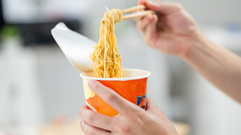 Hands holding chopsticks, lifting ramen noodles out of a cup.
