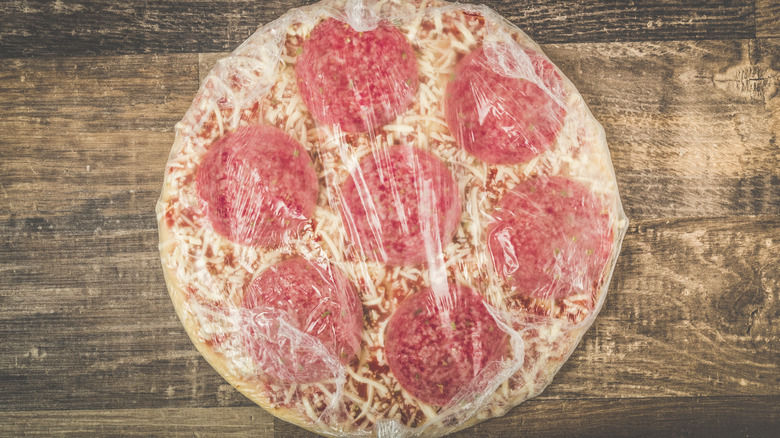 Frozen peperoni pizza wrapped in plastic wrap on a table.