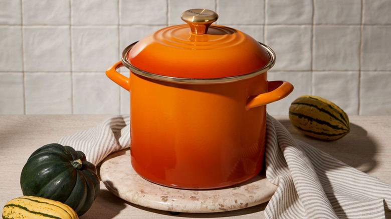 The Le Creuset Stockpot in Persimmon with Gold Hardware against a white background with gourds and striped cloth.