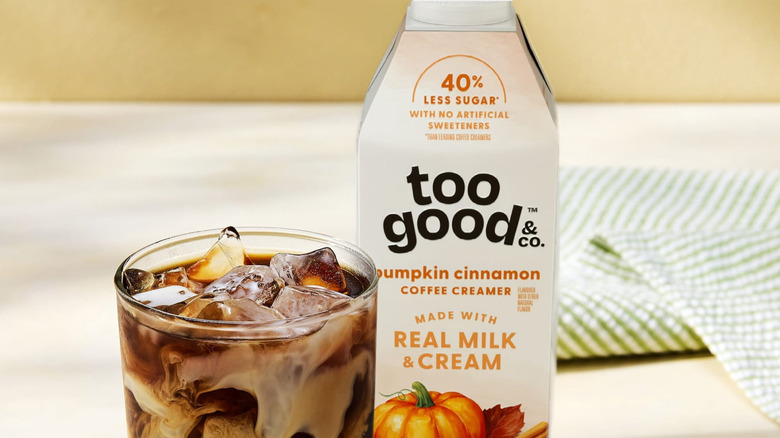 A carton of Too Good & Co. Pumpkin Cinnamon Coffee Creamer on a table next to a glass of iced coffee