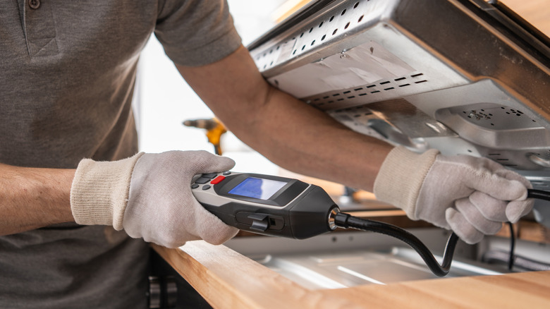 A person in a gray shirt and white gloves handling the underside of a gas stovetop while holding a meter.