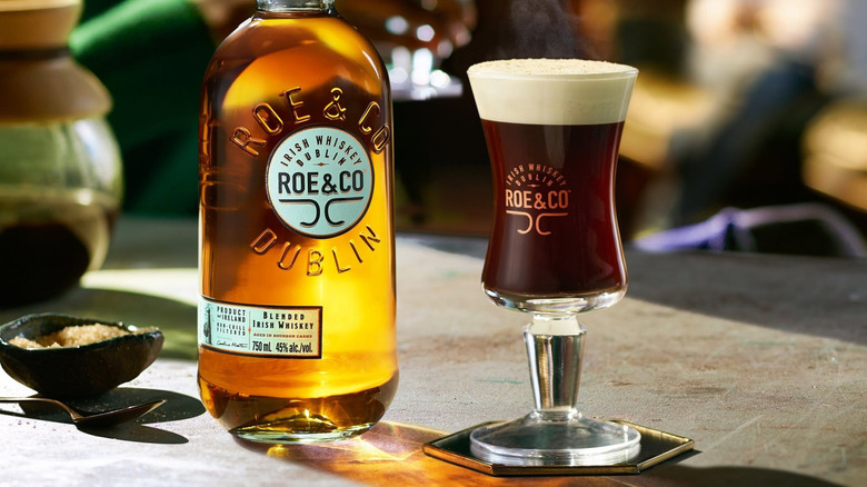 A bottle of Roe & Co Irish Whiskey on a table next to an Irish coffee in a branded glass