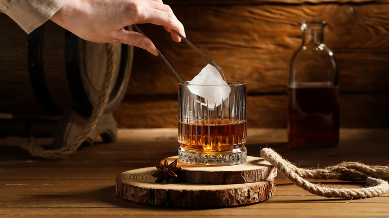 A large ice cube being placed by a person with tongs into a filled whiskey glass in a rustic setting