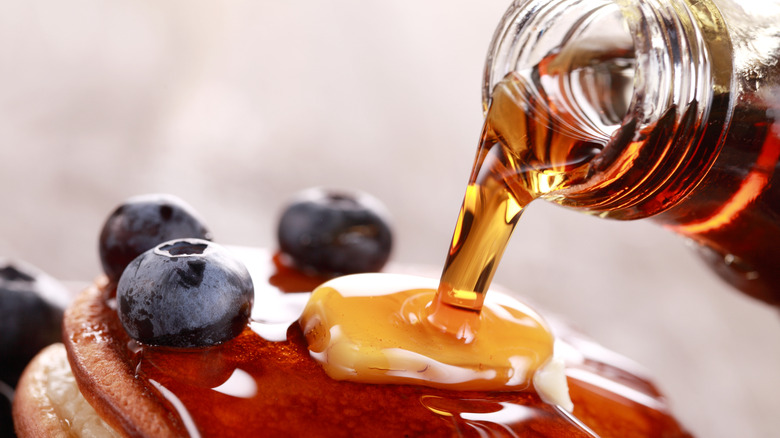 A closeup of a glass bottle of maple syrup pouring over a stack of pancakes with fruit and butter.