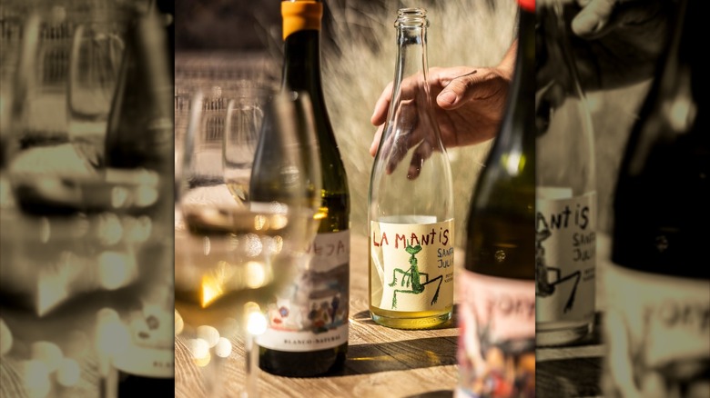 A hand reaches for a white wine bottle with a praying mantis on the label.