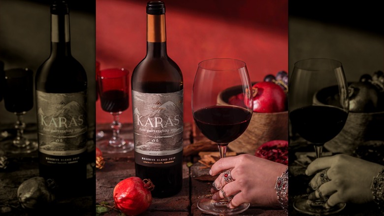 A woman's bejeweled hand holds a red wine glass with Karas wine bottle nearby