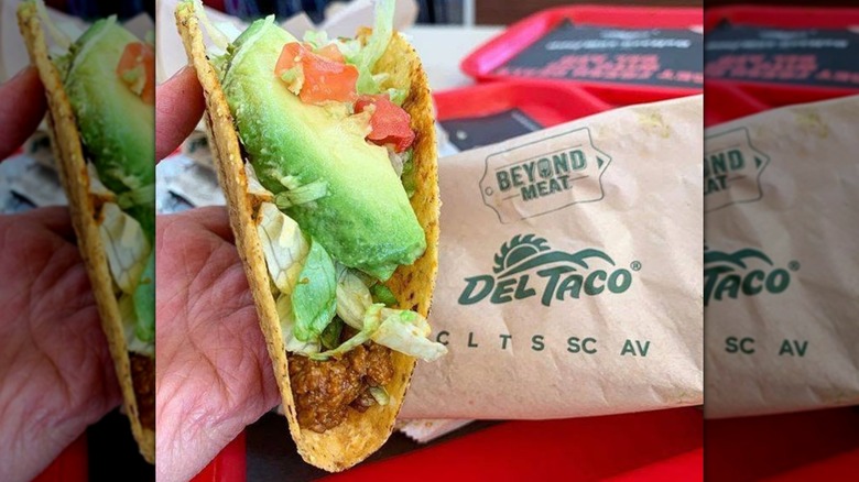 Close up of hand holding taco in front of Beyond Meat Del Taco packaging, mirrored image