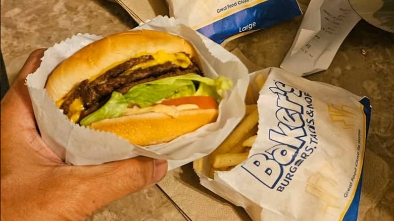 A hand holding a cheeseburger with a bag of Baker's fries on a table.