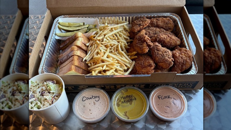 Large container of bread, pickles, fries, fried chicken, next to large container of coleslaw and three sauce containers
